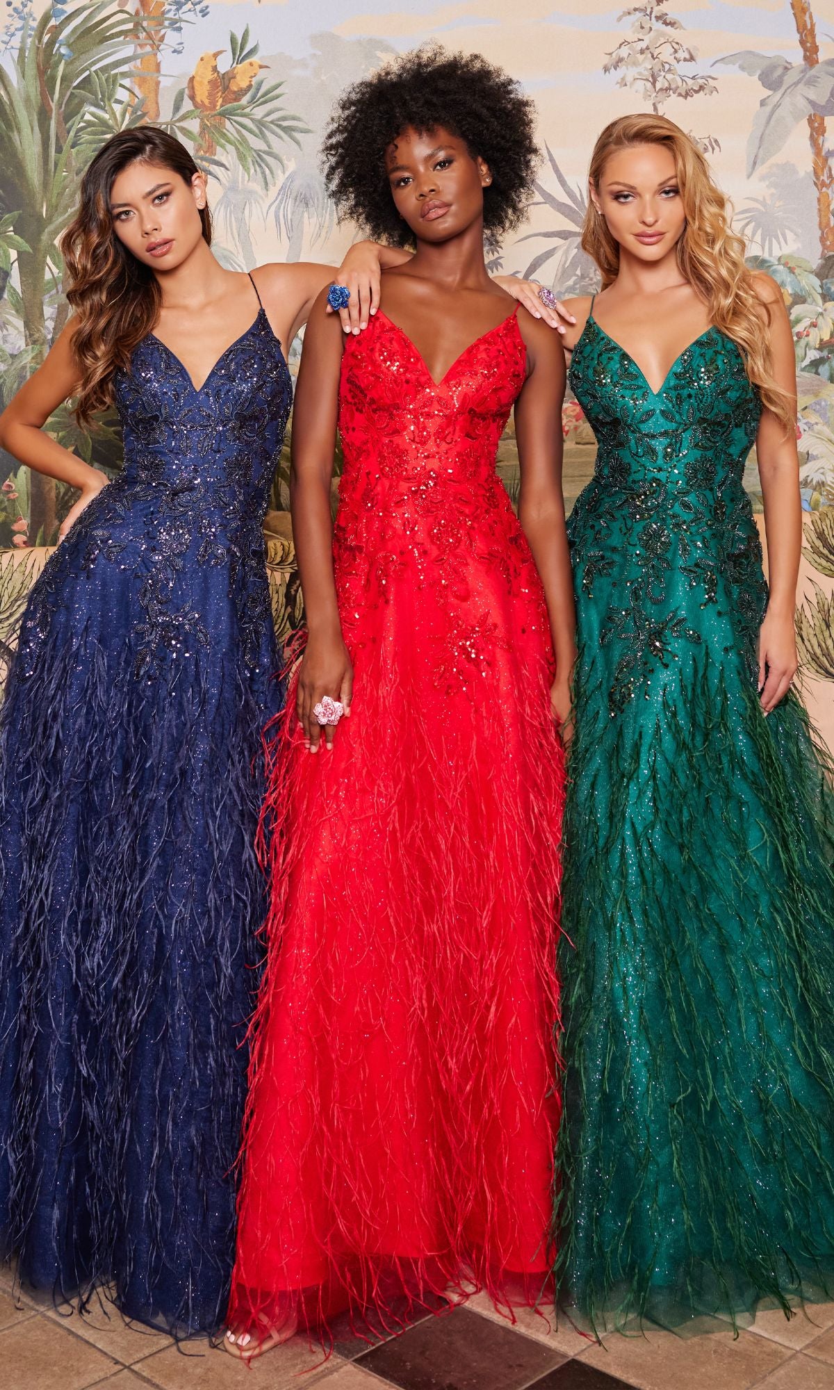 Three women wear the Colette CL2044 Feather Prom Ball Gown in blue, red, and green, featuring sequin and feather details, as they pose side by side against a decorated background.